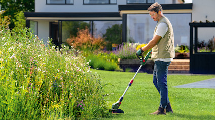 En person klipper gräs med en elektrisk trimmer i en trädgård framför ett modernt hus.