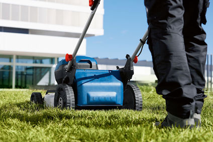 Person klipper gräset med en blå gräsklippare på en gräsmatta nära en modern byggnad.