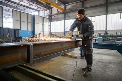 Person i arbetskläder slipar en stor metallbalk i en industriverkstad, gnistor flyger.
