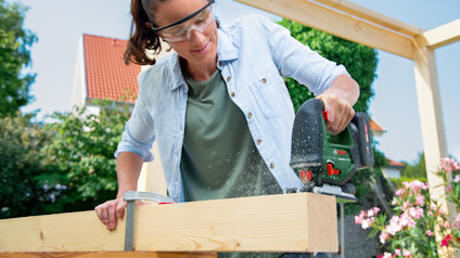 Person som sågar en träbjälke utomhus med en elektrisk såg, bär skyddsglasögon.