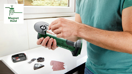 En person håller en grön vinkelslip nära ett fönster. Bredvid finns sandpapper och maskintillbehör. Text: "Magnet Hold".