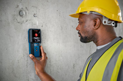 Person med gul hjälm och väst använder digital väggskanner mot en grå betongvägg.