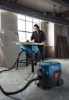 En person arbetar vid ett arbetsbord med blå verktyg och dammsugare i en industrilokal.