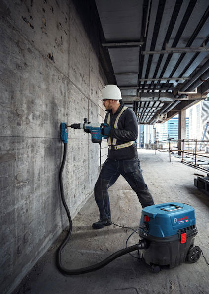 Person med hjälm använder borrverktyg på betongvägg i byggnad under konstruktion. Maskinen ansluten till dammsugare.