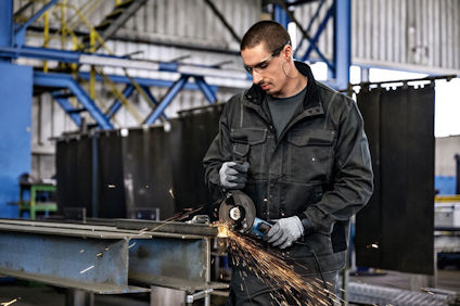 Person slipar metall med vinkelslip i fabrik, gnistor flyger. Bär skyddsglasögon och arbetskläder.