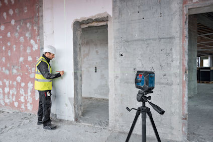 Byggarbetare i gul väst använder mätverktyg på betongvägg; stativ med laserinstrument i förgrunden.