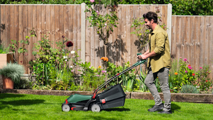 Man klipper gräsmatta med en grön gräsklippare i en trädgård med trästaket och blommor i bakgrunden.