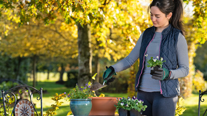 Kvinna i trädgård iklädd handskar planterar blommor i krukor på ett bord. Bakgrunden är träd med höstlöv.
