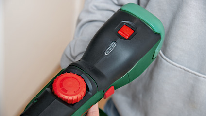 Person som håller i en grön och svart borrmaskin med rött reglage, iklädd grå tröja.
