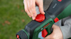 Person justerar en röd knapp på en grön och svart Bosch verktyg med gräs i bakgrunden.