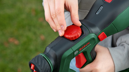 Person justerar en röd knapp på en grön och svart Bosch verktyg med gräs i bakgrunden.