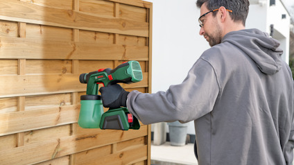 Person i grå tröja och handskar använder grön och svart färgspruta på träpanel.