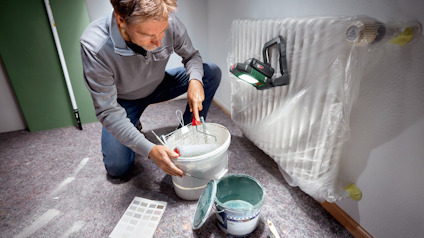 Person målar vägg, använder pensel i målarburk med plast på element, färgprover bredvid.