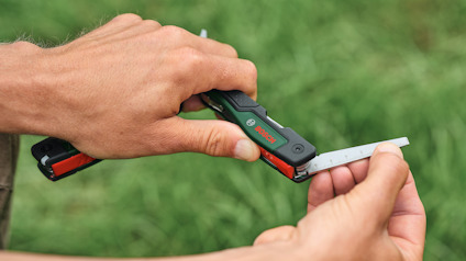 Händer som håller ett Bosch multiverktyg med ett utfällt metallblad mot en grön bakgrund.