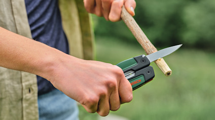 Person använder en multi-tool kniv för att vässa en pinne utomhus.