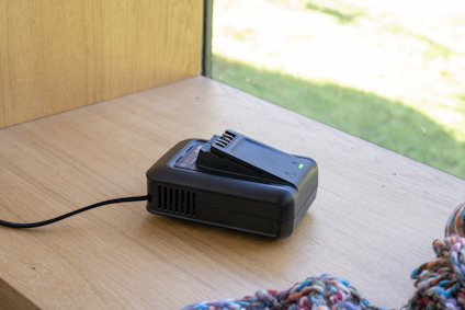 Batteriladdare för laddverktyg på träbord vid fönster, med stickad filt i förgrunden.