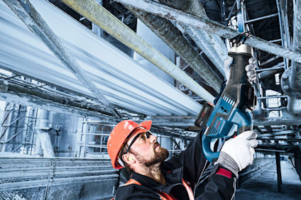 Person med orange hjälm använder en blå elektrisk tigersåg på metallrör i industriell miljö.