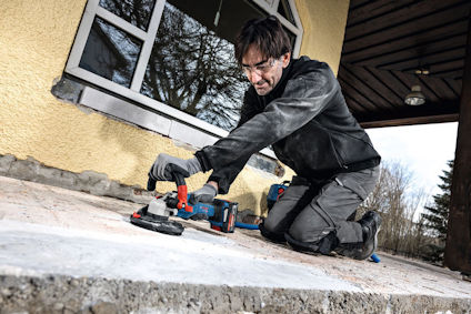 Person som arbetar med en slipmaskin på en betongyta utanför ett gult hus.