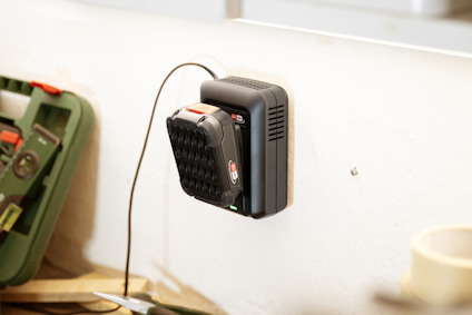 Batteriladdare monterad på vägg, laddar ett svart och rött batteri. Verktygslåda och tejprulle syns i bakgrunden.