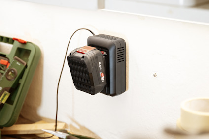 Svart och rött laddningsbart batteri anslutet till en väggladdare. Verktygslåda syns suddigt i bakgrunden.
