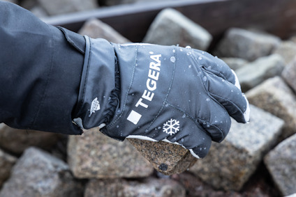 Hand med svart handske, märkt "TEGERA", håller en sten bland andra stenar. Handsken har ett snöflingemärke.