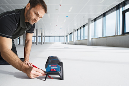 Man mäter ett golv med en Bosch-laseravståndsmätare i ett stort, tomt rum med fönster och vita takplattor.