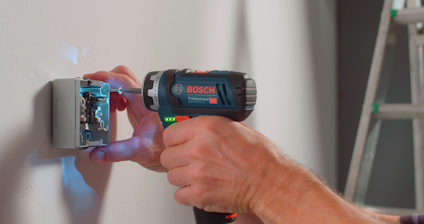 Person använder en Bosch borrmaskin för att installera eller justera en elektrisk enhet på en vägg.梯子 syns i bakgrunden.