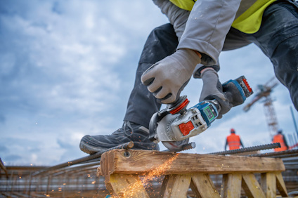 Person som skär genom armeringsjärn med en vinkelslip. Bakgrund med suddiga byggarbetare och kranar mot molnig himmel.