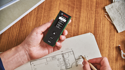Hand håller en Bosch laseravståndsmätare över ett ark med arkitektskisser.
