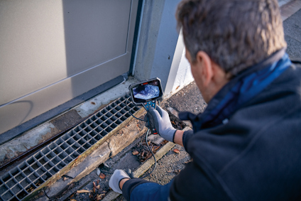 Person inspects a drainage grate and door frame using a handheld inspection camera.