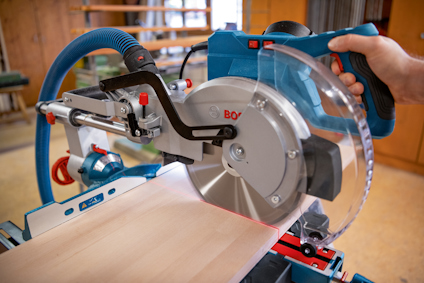 En man använder en elektrisk kapsåg med blått och rött hölje för att såga en träplanka.