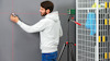En person i vit tröja använder en laser för att mäta en grå vägg, med en stativmonterad enhet intill.