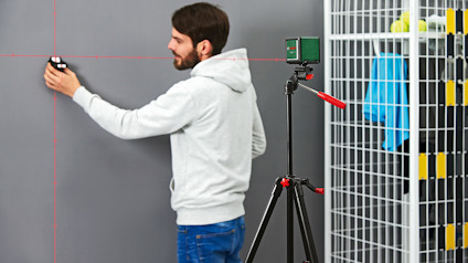 En person i vit tröja använder en laser för att mäta en grå vägg, med en stativmonterad enhet intill.