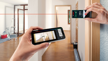 En handenhet mäter avstånd i ett rum med hjälp av en laser, sett genom en smartphone-skärm.