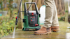 Man med högtryckstvätt rengör en träyta utomhus, beige byxor och bruna kängor.