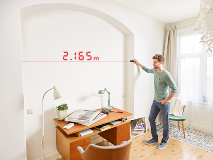 En person använder en laseravståndsmätare för att mäta en vägg, visar 2,165 m. En skrivbordslampa och böcker på skrivbordet.