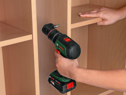 En person använder en grön och svart Bosch borrmaskin på en trähylla.