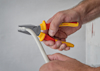 Två händer håller en tång med röd-gult handtag, märkt "BOSCH", böjer en vit kabel.