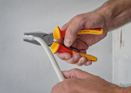Två händer håller en tång med röd-gult handtag, märkt "BOSCH", böjer en vit kabel.