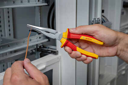 En hand håller en Bosch-avbitartång och klipper en tunn tråd framför en elcentral.