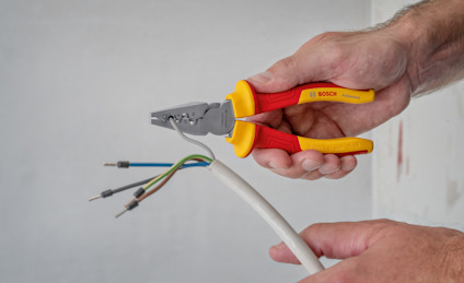 Person håller i en kabelskalare och klipper en kabel med färgade ledningar: brun, blå, gulgrön.