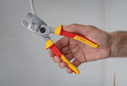 En person håller i en kabelsax med röd-gult handtag märkt "Bosch", som klipper en kabel.
