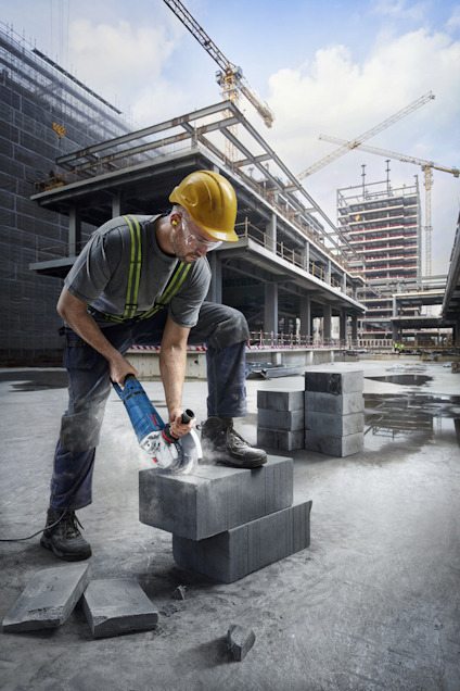 Arbetare med gul hjälm och hörselskydd skär betongblock med en elektrisk såg på byggarbetsplats, med kranar i bakgrunden.
