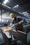 Arbetare i hjälm och skyddsmask slipar metallbalk med vinkelslip i industriell verkstad.