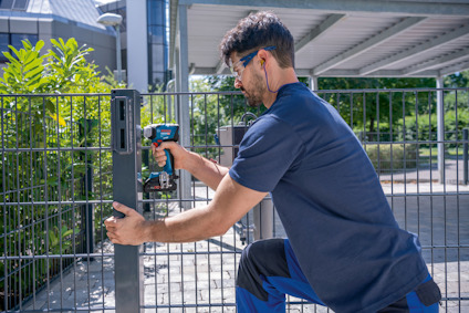 En person i mörka kläder installerar en låsbox på ett metallstängsel med en skruvdragare utomhus.