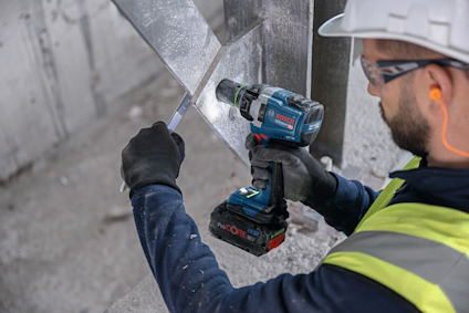 Person i skyddsväst och hjälm använder en blå Bosch borrmaskin på metall.