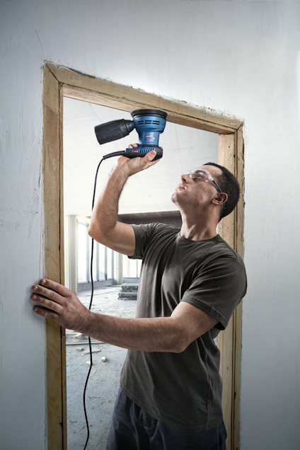 Man using an electric sander on a wooden door frame, wearing safety goggles and ear protection.