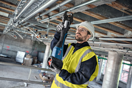 En person i skyddshjälm och gul väst använder en elektrisk skruvdragare på rör i taket av en byggarbetsplats.