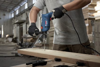En person borrar i ett träplank med en elektrisk borrmaskin i en industrimiljö.