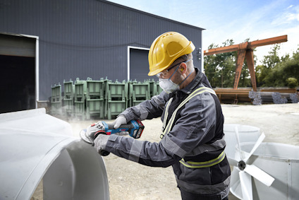 En person bär skyddsutrutning och slipar en stor, vit metallstruktur utomhus nära en industribyggnad.
